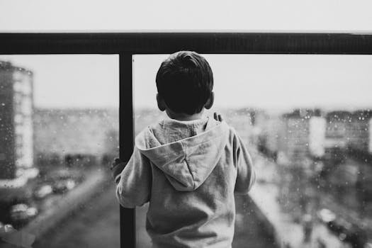 A young boy in a hoodie looks out a rainy window, creating a moody black-and-white scene.